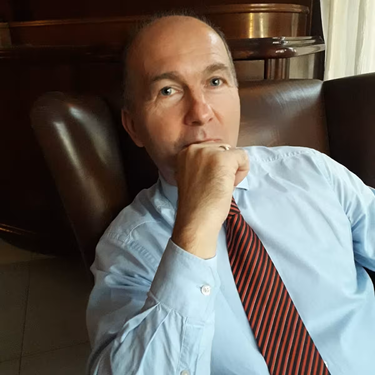 Man in blue shirt and red striped tie resting chin on fist while seated in leather chair.